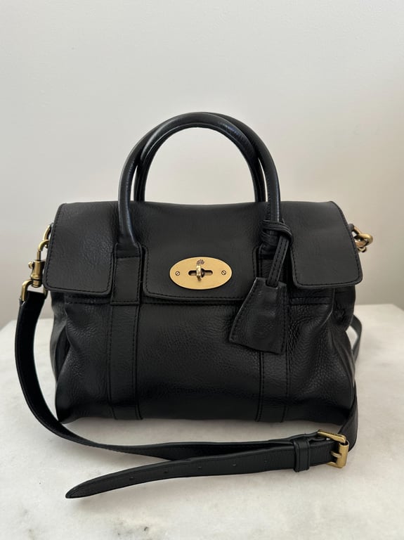 Sort lædertaske med hank og aftagelig skulderrem, foran med gyldent metalspænde, placeret på hvid overflade.