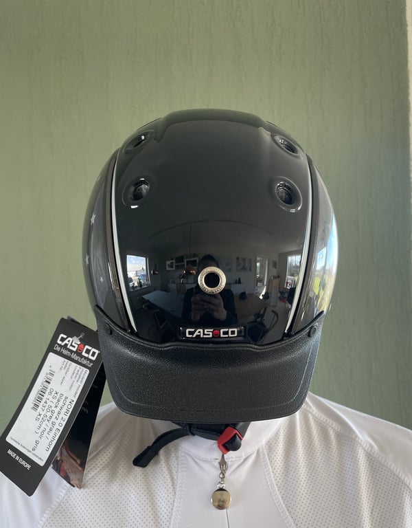 Sort cykelhjelm med reflekterende overflade fra mærket Casco, monteret på en hvid skjortekrop mod en beige væg.