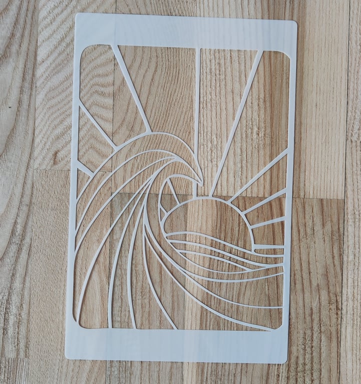 Hvid papirskåret stencil med motiv af en bølge og solstråler mod en solnedgang på en træbordplade.