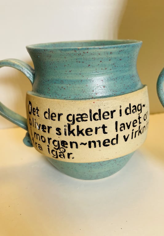 Blåligt keramikkrus med beige bånd og dansk tekst, der siger: "Det der gælder i dag bliver sikkert lavet om i morgen - med virkning fra i går."