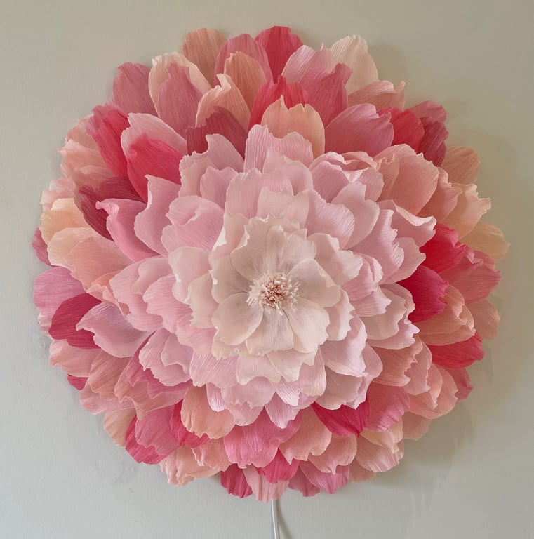 Stor rund vægdekoration lavet af lag-på-lag papirblomster i forskellige nuancer af lyserød og rød.