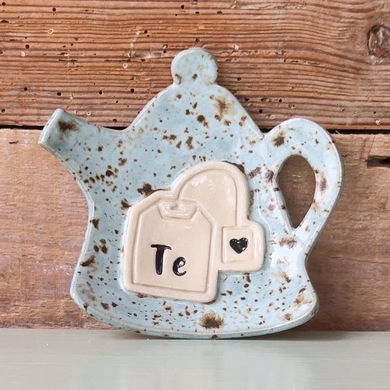 Keramisk te-kande formet lille fad med en tepose og teksten "Te" på forsiden, placeret foran en træbaggrund.