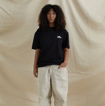 Kvinde med krøllet hår iført sort t-shirt og lyse bukser står foran en beige baggrund.