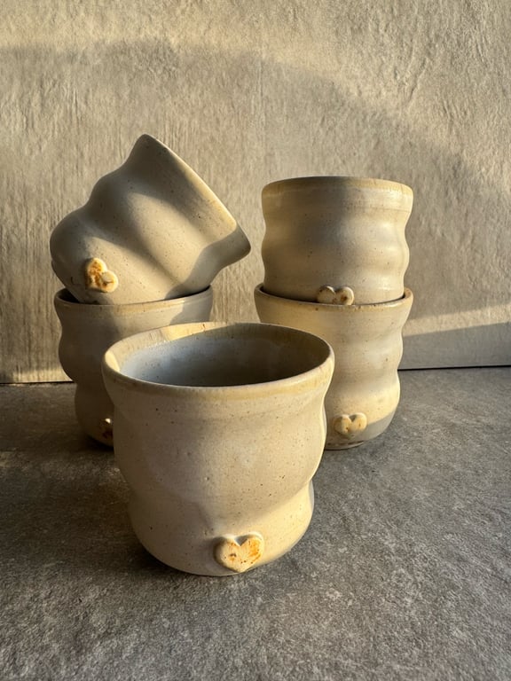 Billedet viser seks lyse keramikkopper med bølgede former, arrangeret i to bunker mod en beige væg. Hver kop har en lille hjerteformet dekoration i bunden.