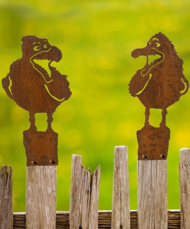 To rustne metalfigurer formet som sjove, grinende fugle er fastgjort på toppen af gamle træhegnspæle mod en sløret, grøn baggrund.