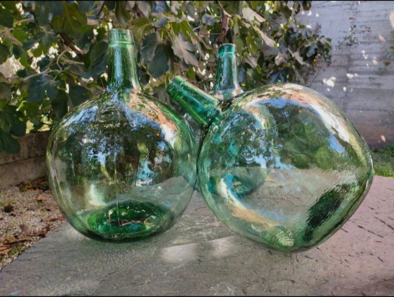 Tre store, grønne, runde glasflasker placeret udendørs på en stenoverflade med grønt løv i baggrunden. (2)