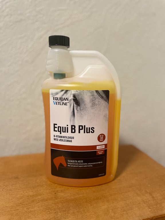 Flaske med Equi B Plus B-vitamintilskud til heste med æblesmag, 1 liter, stående på et bord.