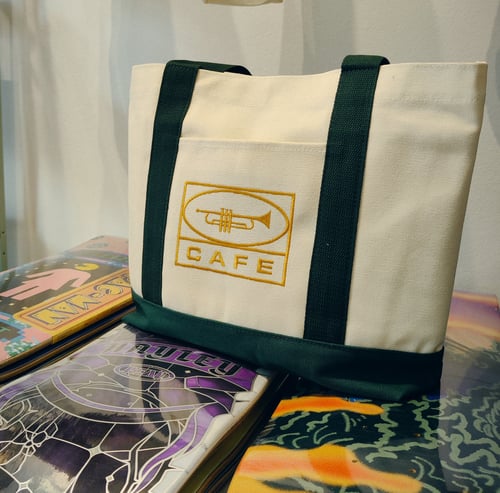 Hvid og grøn stoftaske med et gult trompetlogo og teksten "CAFE", placeret på stakke af farverige vinylpladeomslag. (2)