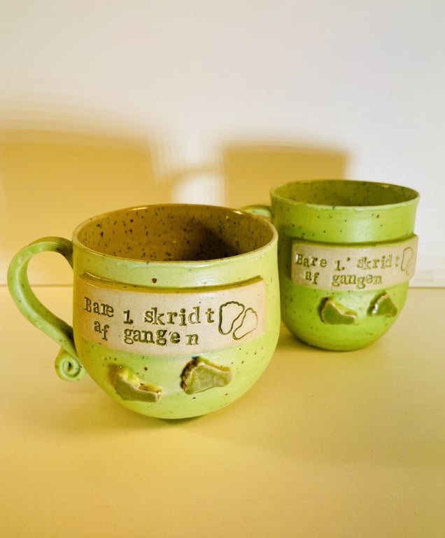 To grønne keramikkrus med teksten "Bare 1 skridt ad gangen" og små fodaftryk som dekoration.