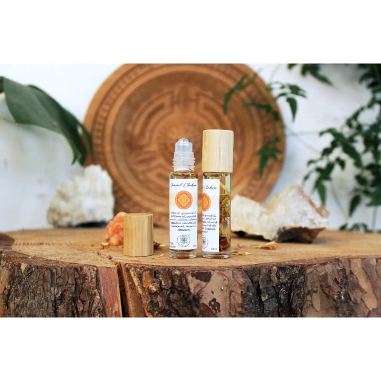 To små glasflasker med rulleapplikator og trælåg stående på et råt træstykke, omgivet af krystaller og grønne blade i baggrunden. (4)