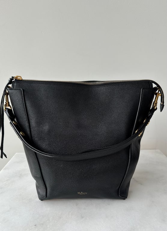 Sort lædertaske med gyldne metaldetaljer og justerbar skulderrem, stående på hvid overflade foran en lys væg.