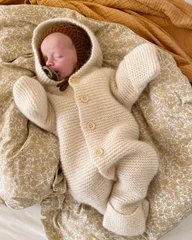 En sovende baby iført en strikket, beige heldragt med hætte og vanter, liggende på et smukt mønstret tæppe. (2)