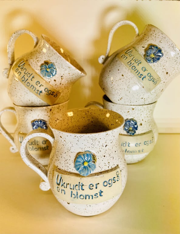 Keramiske, beige kaffekrus med blå blomst og teksten "Ukrudt er også en blomst" skrevet på dem.