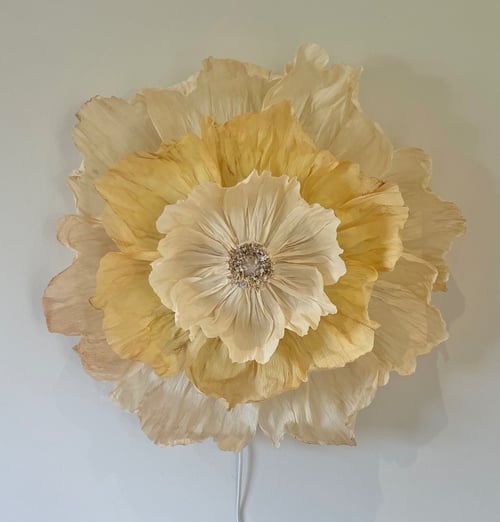 Stor kunstig blomst med flere lag af beige og lysegule kronblade monteret på en lysegrå væg. (1)