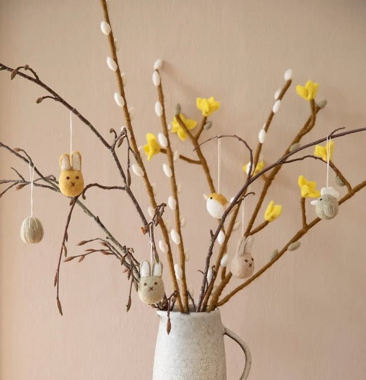 En lysegrå keramikkande med forårsgrene pyntet med gule blomster og ægformede ophæng i filt, herunder kanin- og fuglefigurer.