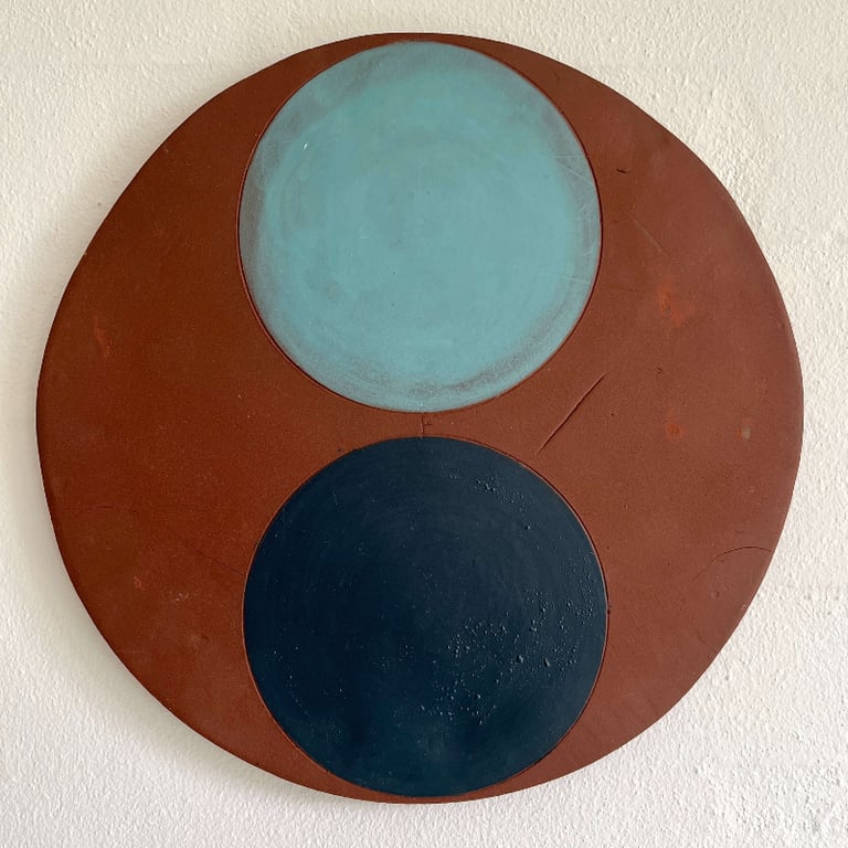 Abstrakt rundt maleri med to store cirkler i blå og sort på en rustfarvet baggrund, monteret på en hvid væg.