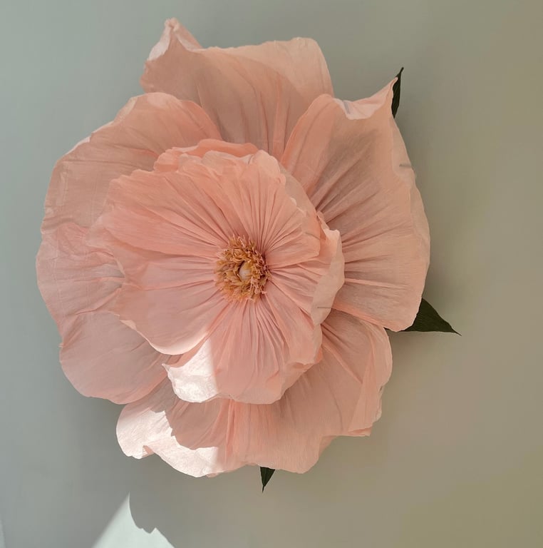 Stor, sart rosa blomsterdekoration med mange folder i papirlignende materiale, monteret på en lys væg.