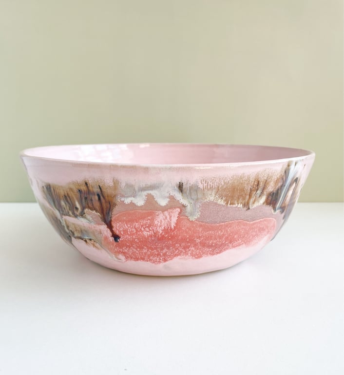 Lyserød keramisk skål med dekorative brune, hvide og sorte glasurdetaljer, placeret på hvid overflade mod en lys beige væg.