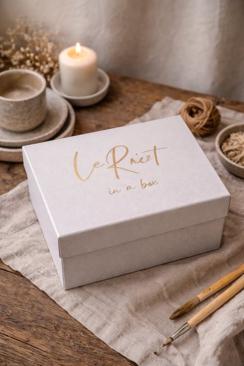 En hvid boks med teksten "Le Rit in a box" i guldskrift, placeret på et lyst stof på et rustikt træbord, omgivet af en tændt lys, en kop med underkop, to pensler og en skål med snor og tørrede planter. (1)
