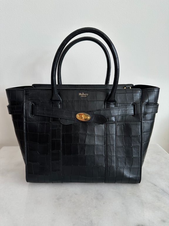 Sort Mulberry håndtaske i croco-præget læder med guldhardware, stående på hvid overflade foran hvid væg.