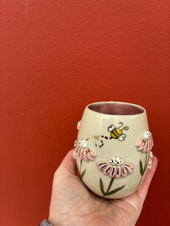 Hånd holder en cremefarvet keramik kop med 3D lyserøde blomster og en malet bi mod en rød baggrund.