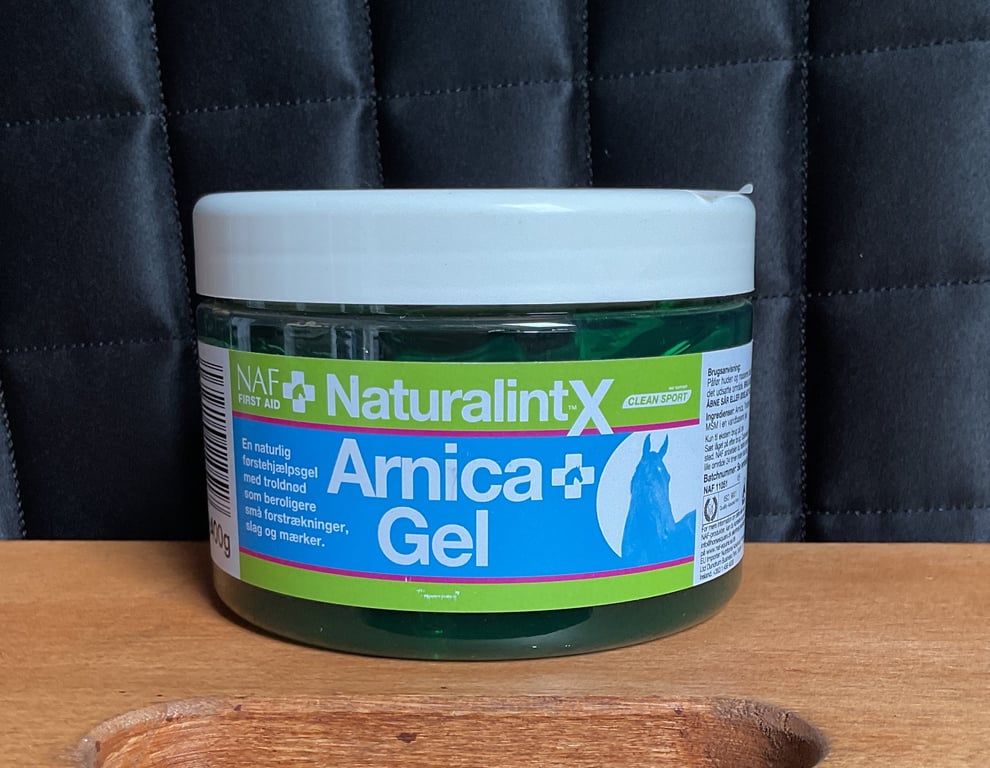 Beholder med NAF Naturalint X Arnica Gel, en naturlig førstehjælpsgel til heste, placeret på et træbord med sort polstret baggrund.