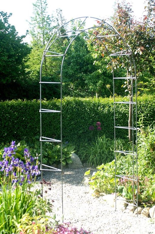 Havebue i metal placeret på grussti omgivet af blomster og grønne hække.