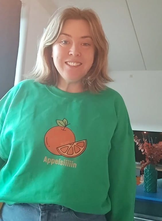 Kvinde med skulderlangt lyst hår iført grøn sweatshirt med illustration af appelsin og teksten "Appelsiiiiin". (3)