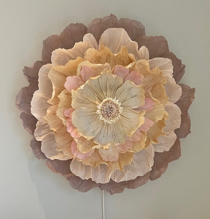 Stor blomst af papir med flere lag i beige, lyserøde og brune nuancer på en lys væg.
