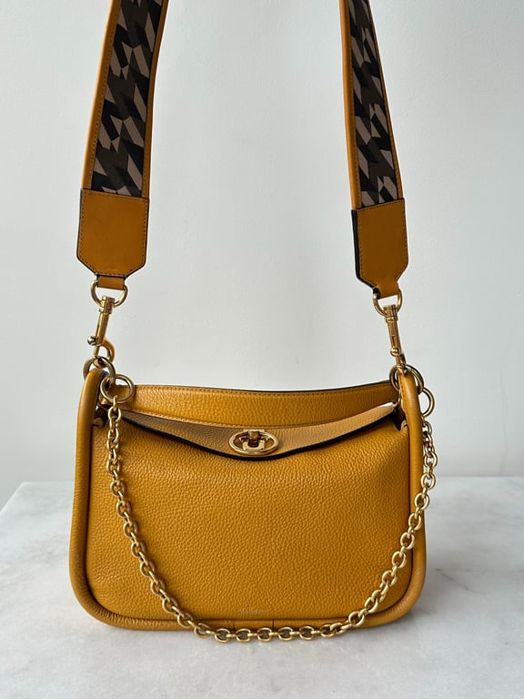 Sennepsgul lædertaske med gylden kæde og bredt skulderrem med sort mønster, sat på hvid overflade foran hvid væg.