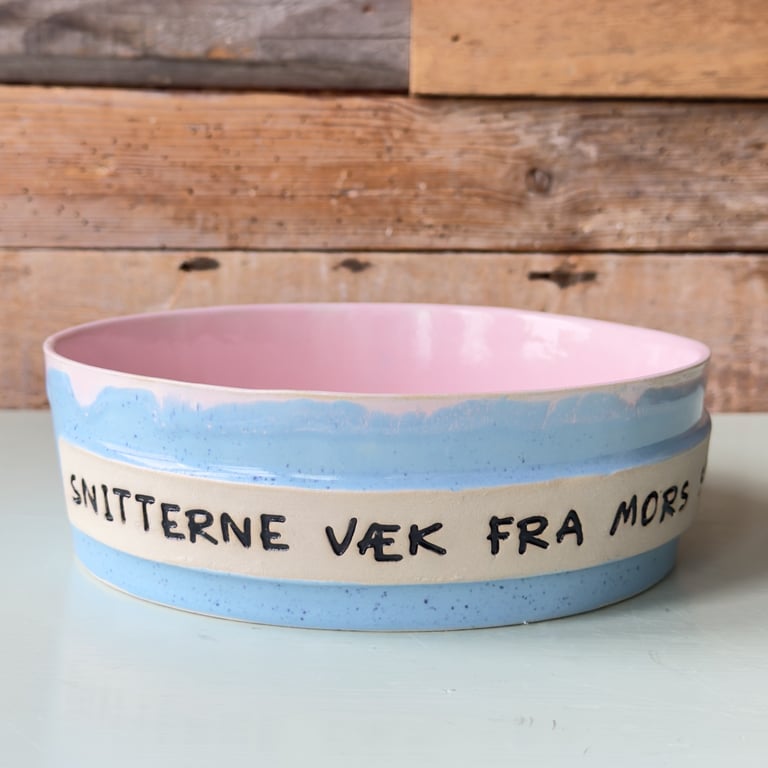 En keramisk skål med en lyseblå yderside og rosa inderside, med teksten "SNITTERNE VÆK FRA MORS" skrevet på siden. Skålen står på en lys overflade foran en væg af genbrugstræ.