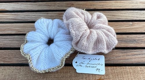 To strikkede scrunchies i hvid og lyserød med gyldent kantbånd, lagt på et træbord, med en pris- og materialelap ved siden af. (3)