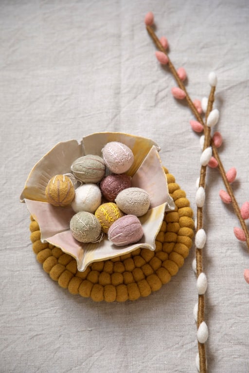 Skål med farverige, broderede påskeæg i pastelfarver placeret på en gul filtunderlag med to grene med hvide og rosa knopper ved siden af på en lys dug.