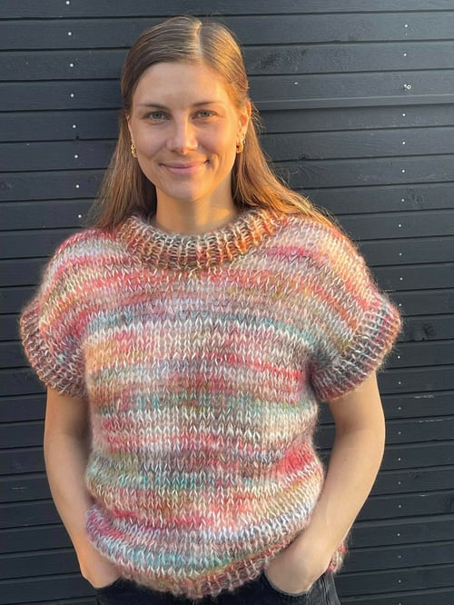Portræt af smilende kvinde med langt lyst hår, iført en kortærmet, flerfarvet strikket sweater foran en sort trævæg. (1)
