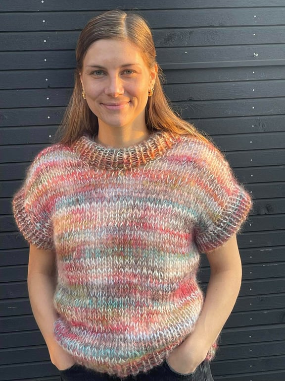 Portræt af smilende kvinde med langt lyst hår, iført en kortærmet, flerfarvet strikket sweater foran en sort trævæg.