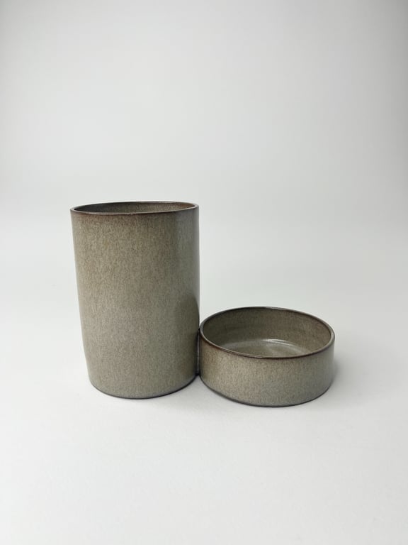 To grå keramiske beholdere med mat finish, en høj og cylindrisk, den anden lav og rund, placeret på hvid overflade.
