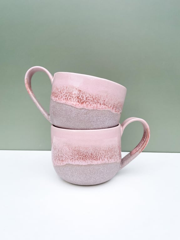 To lyserød og grå keramikkrus stablet mod en grøn væg og hvid overflade.