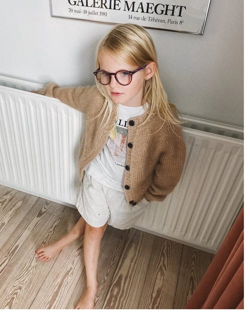 En ung pige med langt blondt hår og briller står afslappet med ryggen mod en radiator og en plakat på væggen bag sig. Hun har en beige strikket cardigan, en hvid t-shirt og lyse shorts på, og hun står barfodet på trægulvet. (4)