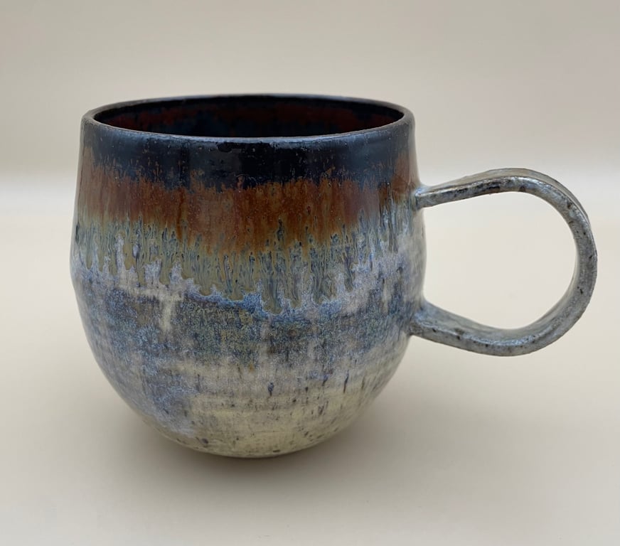 Krus i keramik med glaseret overflade i blå, grå og brunlige nuancer, med rund form og stort håndtag.