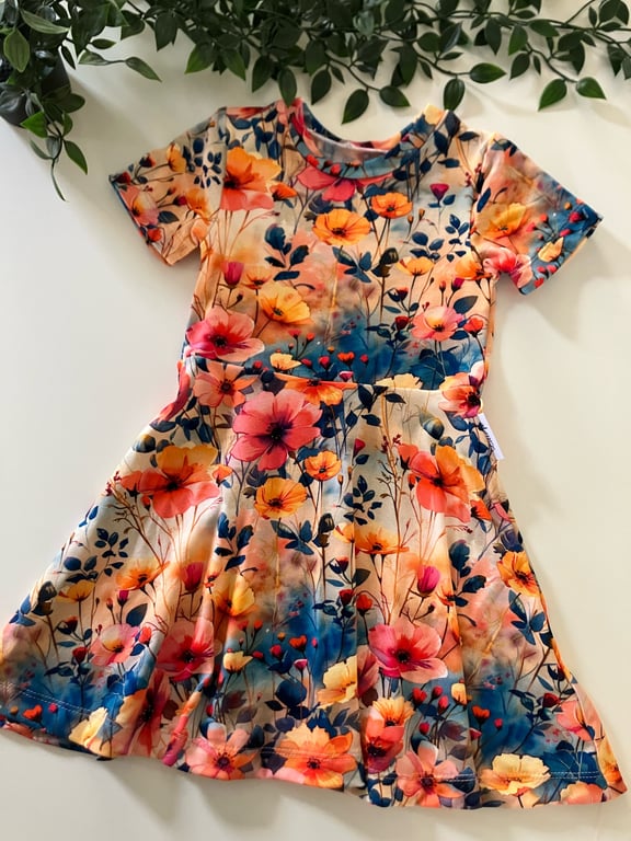 Farverig børnekjole med korte ærmer og blomsterprint i nuancer af orange, pink, gul og blå, lagt fladt på en hvid overflade med grønne blade omkring.