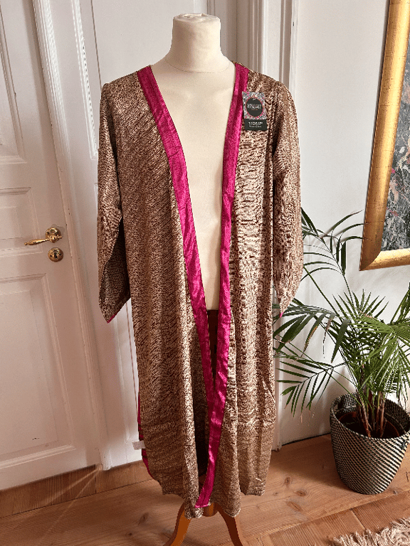 Mannequin med lang kimono i brunligt mønster med pink satin kant, placeret i et lyst rum med trægulv, grøn plante og del af et maleri i guldramme. (2)