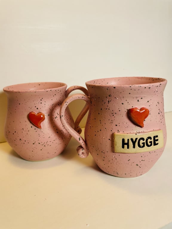 To rosa keramikkrus med sort spættet mønster, røde hjerter og et krus med skilt med teksten "HYGGE".