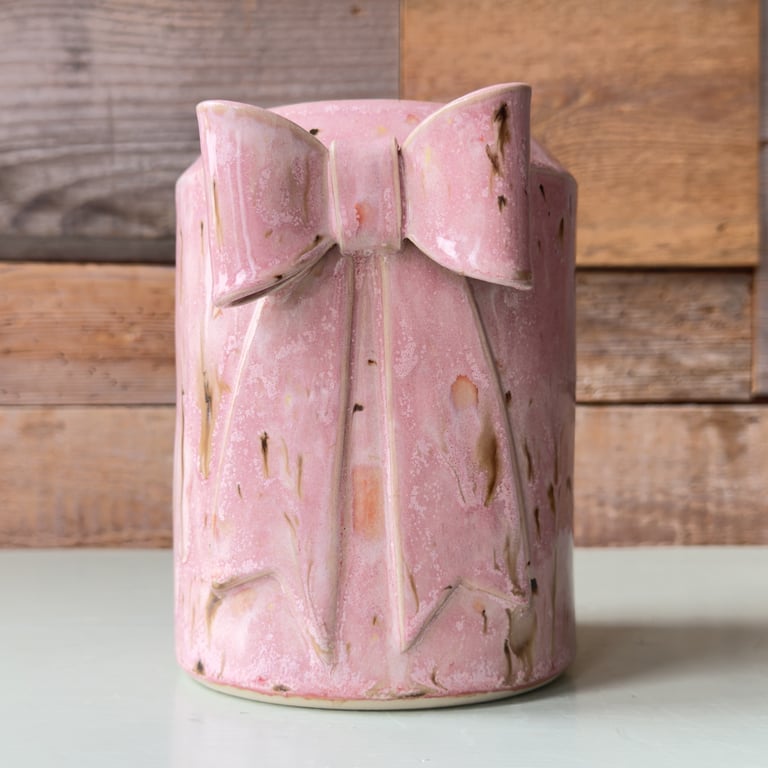 Lyserød keramisk vase med relieff af en sløjfe, placeret på en lysegrå overflade med træpaneler i baggrunden.