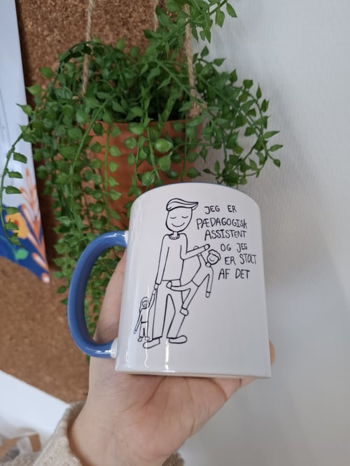 Hånd, der holder en hvid kaffekop med blå hank og tegning af en voksen og to børn samt teksten "Jeg er pædagogisk assistent og jeg er stolt af det", foran en grøn plante og en opslagstavle. (2)
