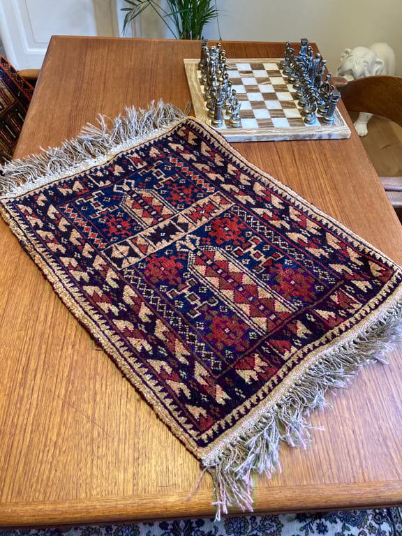 Lille vævet tæppe med geometrisk mønster i blå, rød og beige farver lagt på et træbord, med et skakspil i baggrunden. (2)