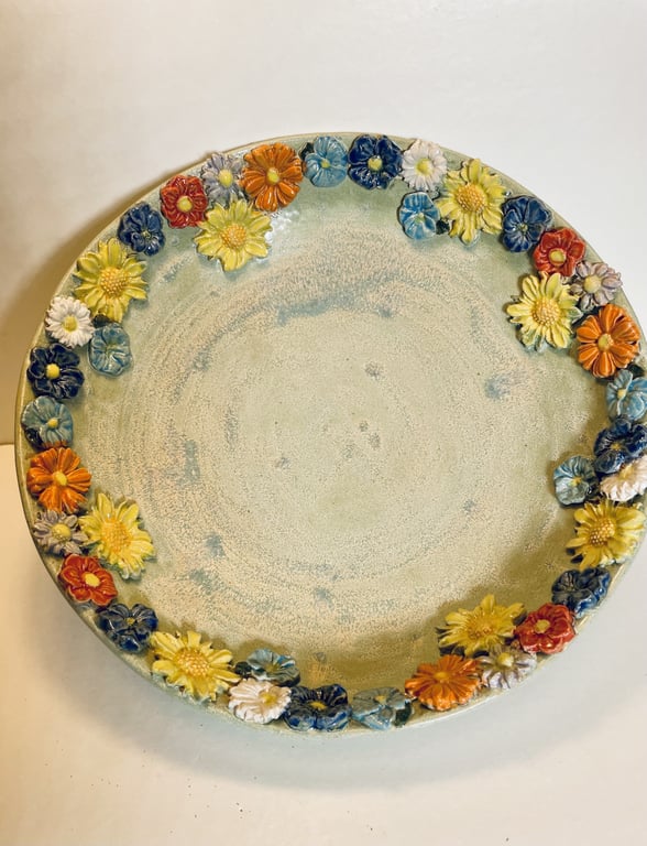 Rund keramik tallerken med blomster i relief i farverne orange, blå, gul og hvid langs kanten.