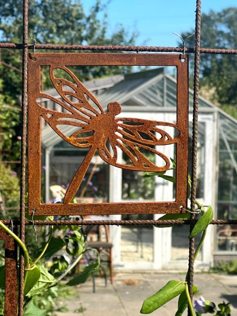 Rustent metaludsmykning af en guldsmed monteret på et metalgitter med et drivhus i baggrunden under en klar blå himmel.