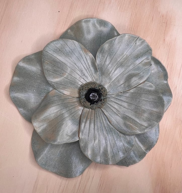 Metalblomst med brede, bølgede kronblade og en sort midte, placeret på en lys træflade.