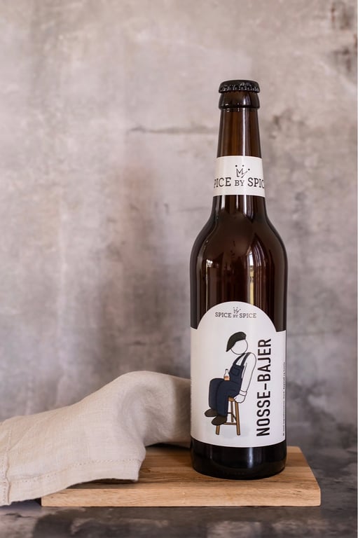 Flaske med øl fra mærket "Nosse-Bajer" stående på en træplade med et lyst stof ved siden af, foran en grå, tekstureret baggrund.