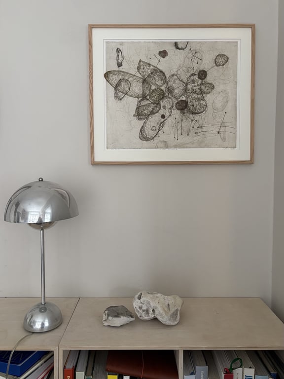 Et minimalistisk hjørne med en sølvfarvet bordlampe, to dekorative sten på et lyst skab og en indrammet abstrakt tegning hængende på en grå væg.